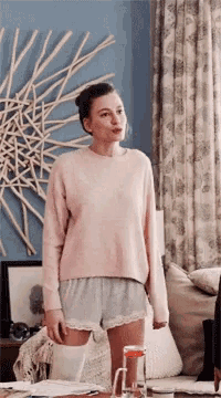 a woman wearing a pink sweater and grey shorts stands in a living room