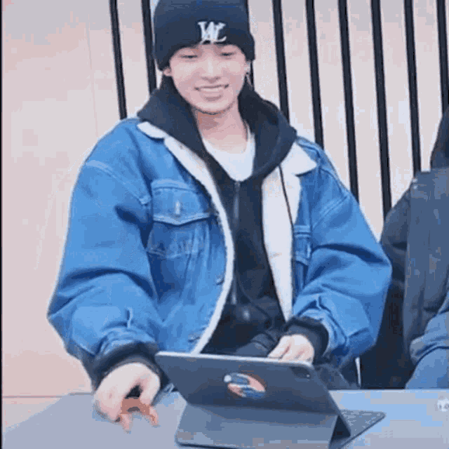 a man wearing a denim jacket and a hat is sitting at a table with a tablet .