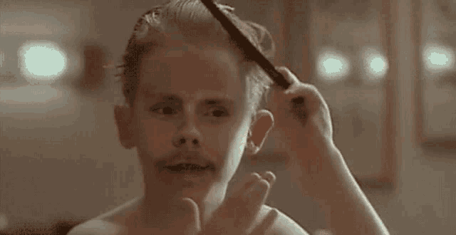 a young boy with a mustache is combing his hair in a bathroom .