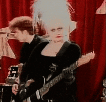 a woman is playing a guitar in front of a red curtain while a man plays drums behind her .