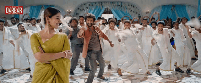 a woman in a yellow saree is standing in front of a crowd of people dancing with the word darbar on the bottom right