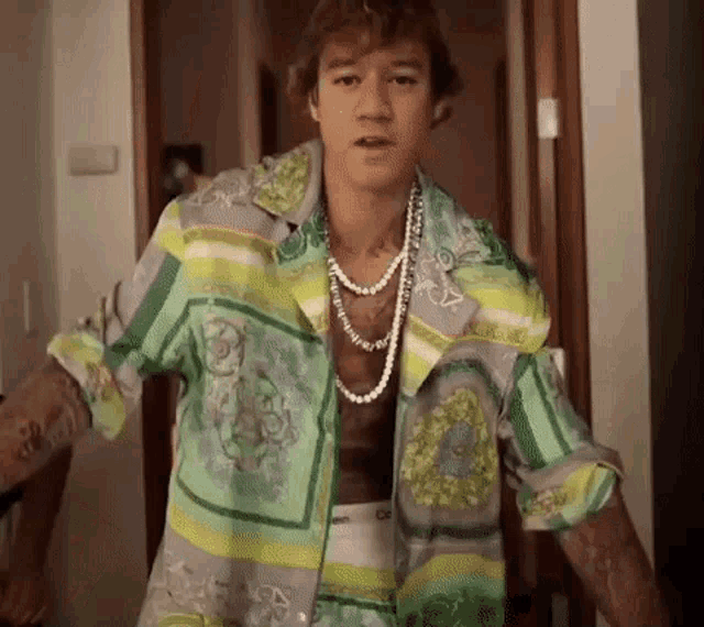 a man wearing a green shirt and a pearl necklace is standing in a hallway .