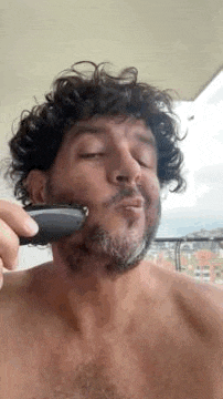 a man without a shirt is shaving his beard with a trimmer .