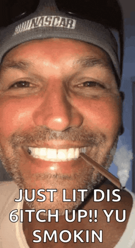 a man wearing a nascar hat is smiling and smoking a cigarette .