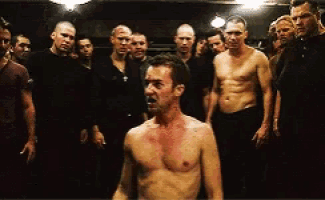 a group of men are standing around a shirtless man in a dark room