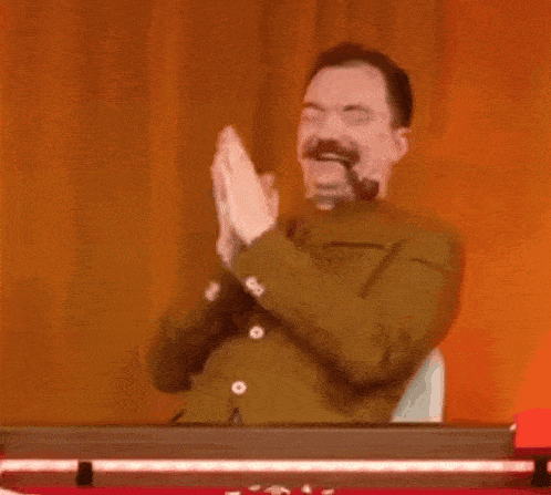 a man with a pipe in his mouth is sitting at a table and clapping his hands .