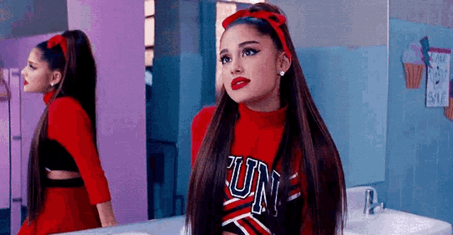 ariana grande is wearing a red cheerleader outfit and standing in front of a bathroom mirror .