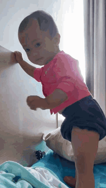 a baby in a pink shirt and blue shorts stands on a bed