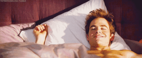 a shirtless young man is laying in bed with his eyes closed and smiling .