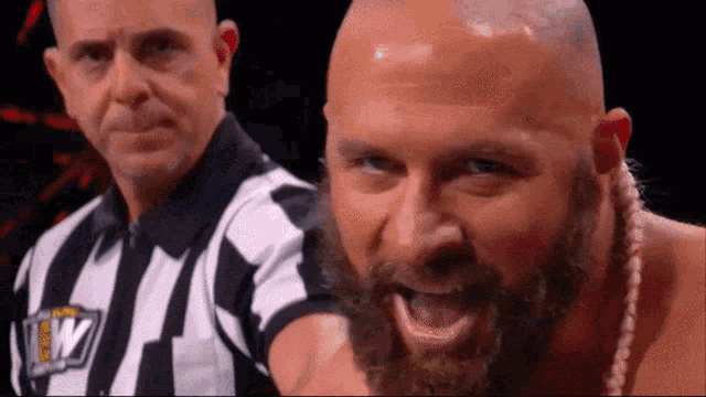 a bald man with a beard is standing next to a referee and making a face .