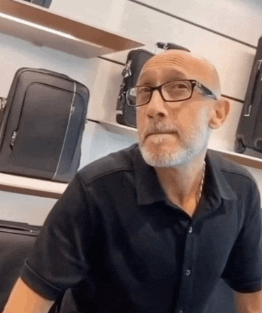 a bald man with glasses and a beard is sitting in front of a display of luggage