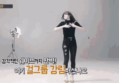 a woman in a black top and black pants is dancing in front of a camera with korean writing on it .