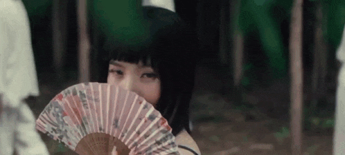 a young woman is holding a fan in front of her face in the woods .
