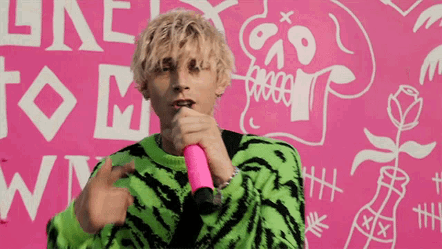 a man in a green and black sweater is holding a pink microphone in front of a pink wall that says " one to one "