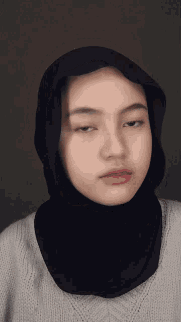 a woman wearing a black hijab and a grey sweater looks at the camera