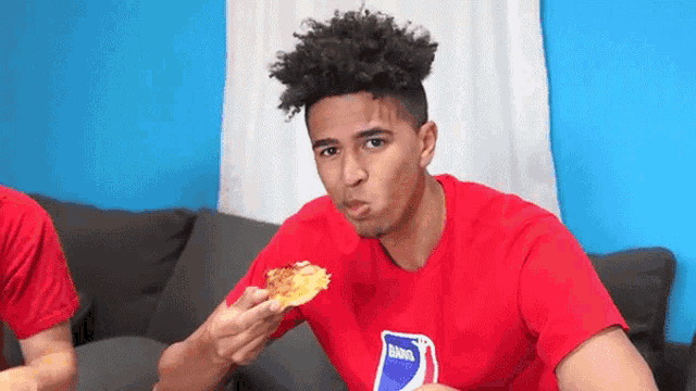 a man in a red shirt is eating a slice of pizza while holding a pepsi card .
