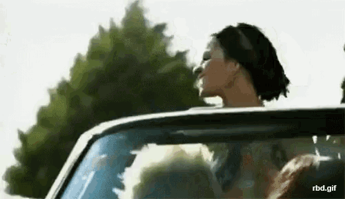 a woman is sitting in a car with her head out the window and trees in the background .