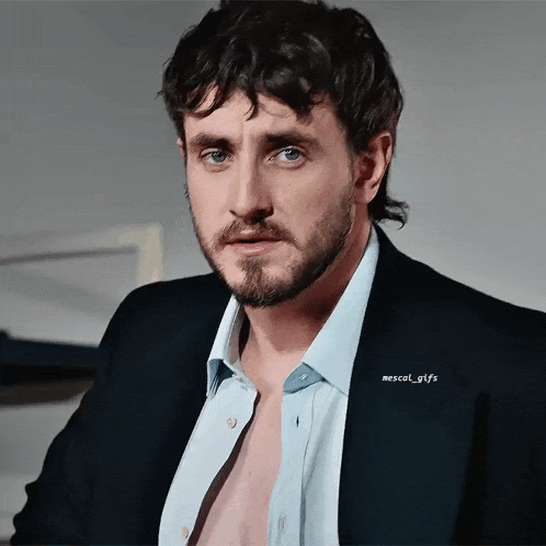 a man with a beard wearing a blue shirt and a black jacket with mescal_gtfs written on it