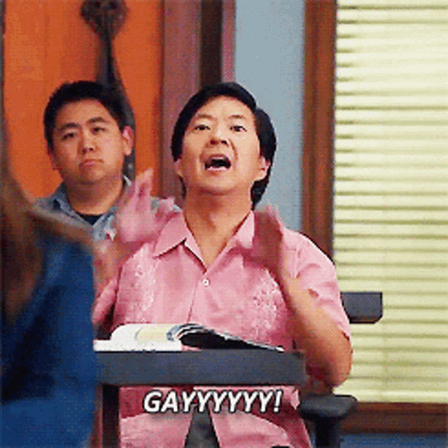 a man in a pink shirt is sitting at a desk and saying gayyyyyy