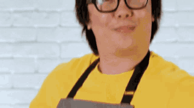 a man wearing glasses and an apron is standing in front of a brick wall .