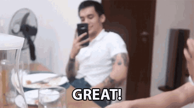 a man is sitting at a table looking at his phone and the word great is visible in the foreground
