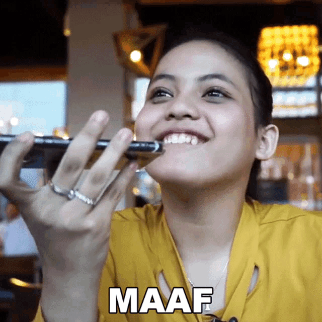 a woman in a yellow shirt is smiling and holding a pen in her mouth with the word maaf below her