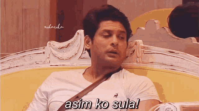 a man in a white shirt is sitting on a yellow couch and says asim ko sula