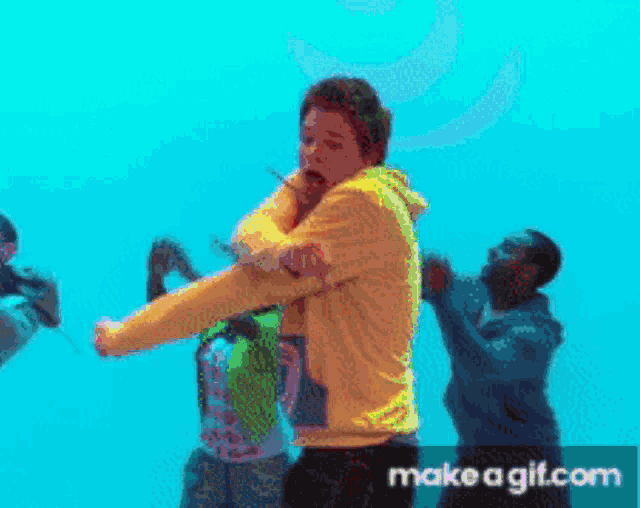 a man in a yellow hoodie is standing in front of a blue background with make a gif.com written on the bottom right