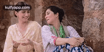 two women are sitting next to each other on a rock and laughing .