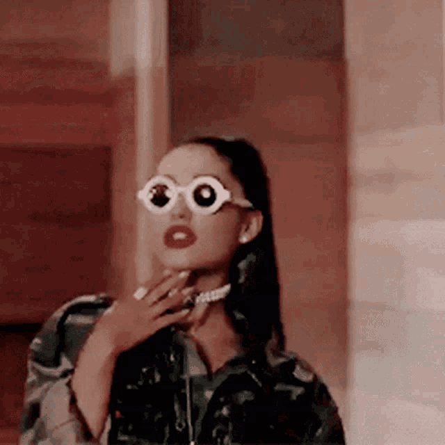 ariana grande is wearing sunglasses and a choker while standing in front of a wall .