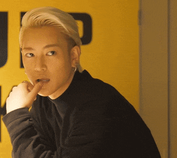 a man with blonde hair and a black turtleneck looks at the camera with his finger in his mouth