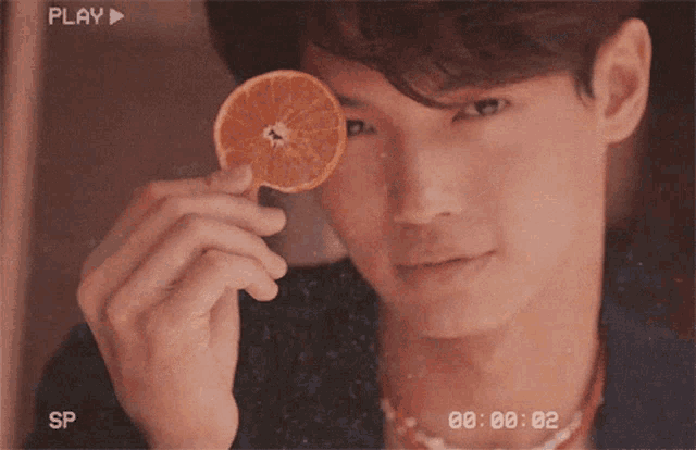 a man is holding a slice of orange in front of his eye and the play button is visible