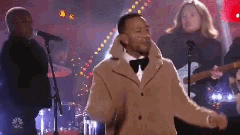 a man in a tan coat and bow tie is singing into a microphone on stage .