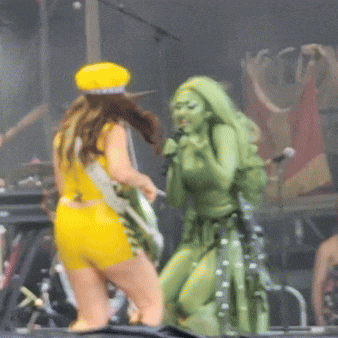 a woman in a yellow outfit is singing into a microphone while another woman in a green outfit stands behind her