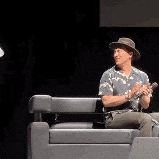 a man wearing a hat is sitting in a chair holding a microphone and clapping .