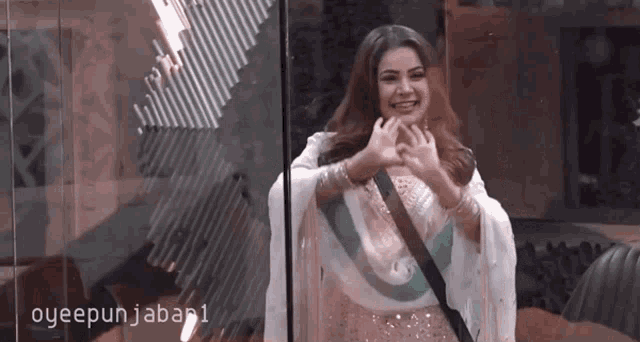 a woman in a white dress is standing in front of a glass wall and making a heart shape with her hands .