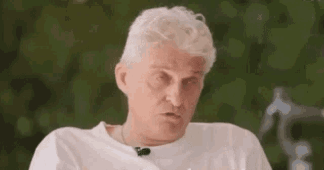 a man with white hair is wearing a white t-shirt and a microphone on his neck .