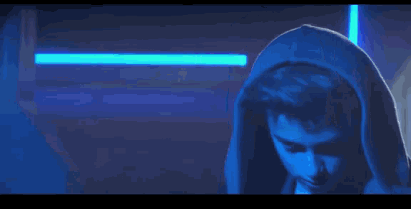 a young man wearing a hoodie is standing in a dark room with blue lights behind him .