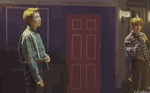 a man in a plaid shirt is singing into a microphone while another man stands in front of a red door .