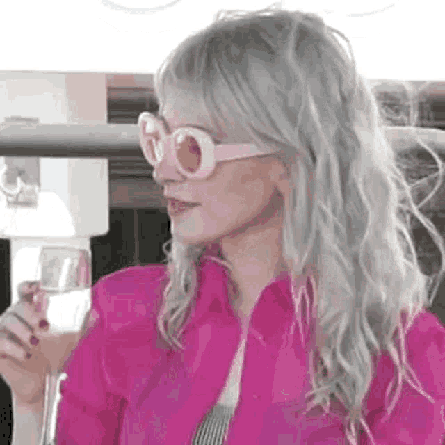 a woman wearing pink sunglasses and a pink jacket is holding a glass of water .