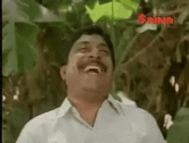 a man with a mustache is laughing with his mouth open in front of a tree .
