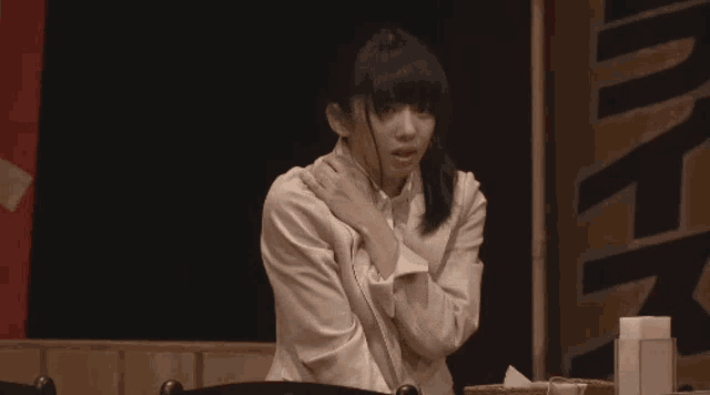 a woman is sitting at a table with her hands on her shoulder and looking at the camera .