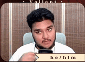 a man with a beard is sitting in a chair holding a cell phone and a sign that says he / him
