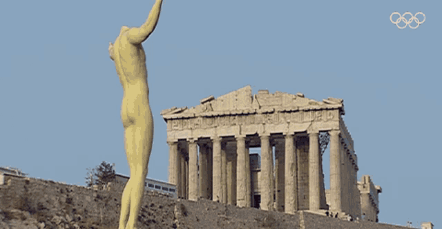 a statue of a naked man stands in front of a building with the olympic rings on the bottom