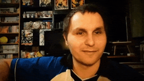 a man in a blue shirt is smiling in front of a shelf full of books