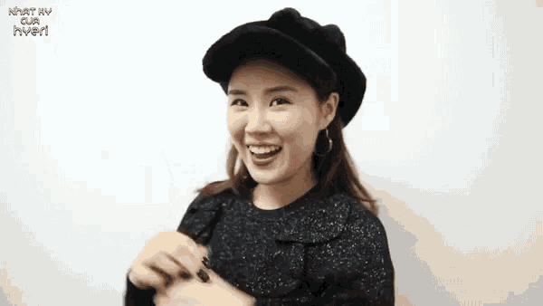 a woman wearing a black hat is smiling in front of a white wall that says nhật hv qua hvem