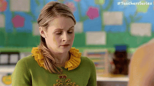 a woman wearing a green sweater and a yellow ruffled collar is standing in a classroom .