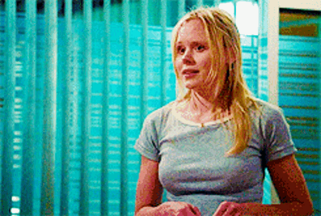 a blonde woman in a grey shirt stands in front of a blue curtain .