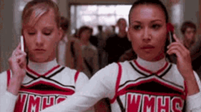 two cheerleaders are talking on their phones in a hallway .