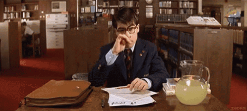 a man in a suit and tie is sitting at a desk in a library looking at papers
